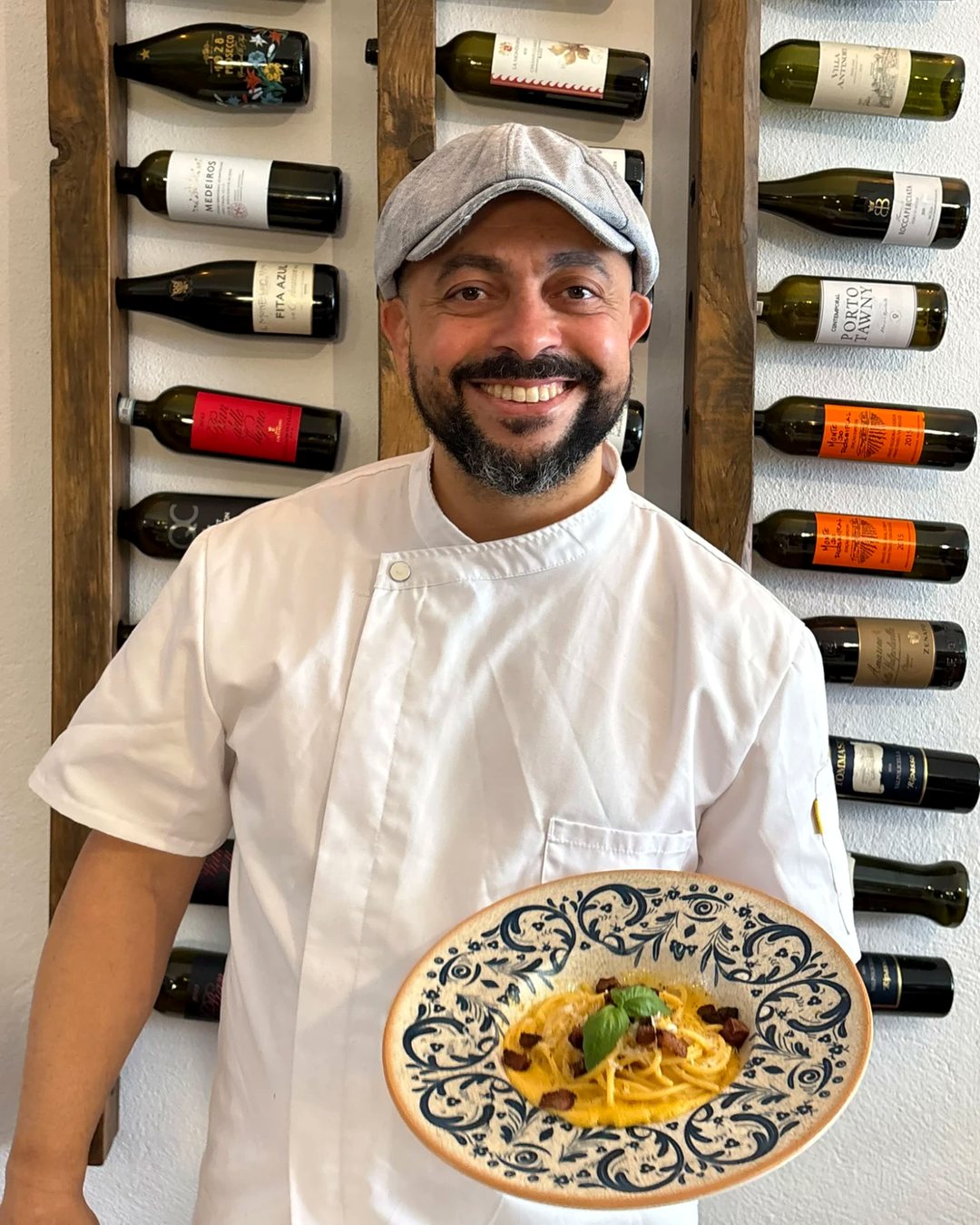 O chef Walei Marcolino, de boina e farda branca, segura um prato de esparguete à carbonara em frente à parede dos vinhos